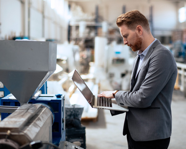 Ein Mann in einem grauen Anzug steht in einer Fabrikhalle und arbeitet an einem Laptop. Er scheint konzentriert zu sein und blickt auf den Bildschirm, während er neben einer großen Maschine steht. Die Fabrik im Hintergrund ist unscharf, zeigt jedoch verschiedene Maschinen und Geräte.