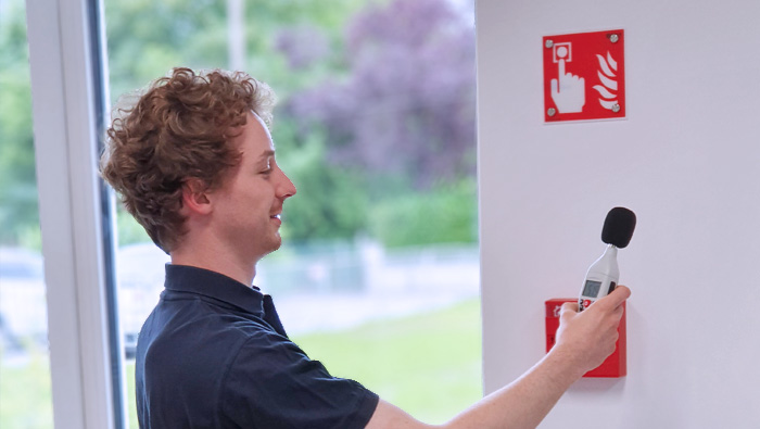 Ein junger Mann in dunkelblauer Kleidung hält ein Schallpegelmessgerät vor eine Wand, auf der ein rotes Brandschutzschild mit einem Symbol für einen Feuermelder angebracht ist. Er scheint die Lautstärke oder Funktion eines Alarmsystems zu testen. Im Hintergrund ist durch ein Fenster eine grüne Außenlandschaft zu sehen. Die Szene deutet auf eine Sicherheitsüberprüfung oder eine Wartung des Alarmsystems hin.