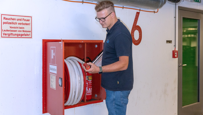 Ein Mann in blauer Arbeitskleidung steht vor einem geöffneten Feuerwehrschrank in einem Gebäude. Im Schrank befinden sich ein aufgerollter Feuerwehrschlauch und ein Feuerlöscher. Der Mann hält den Schlauch und scheint eine Inspektion oder Wartung durchzuführen. An der Wand neben dem Schrank hängt ein Warnschild, das Rauchen und offenes Feuer verbietet. Die Szene deutet auf eine Brandschutzüberprüfung oder Sicherheitsmaßnahme hin.